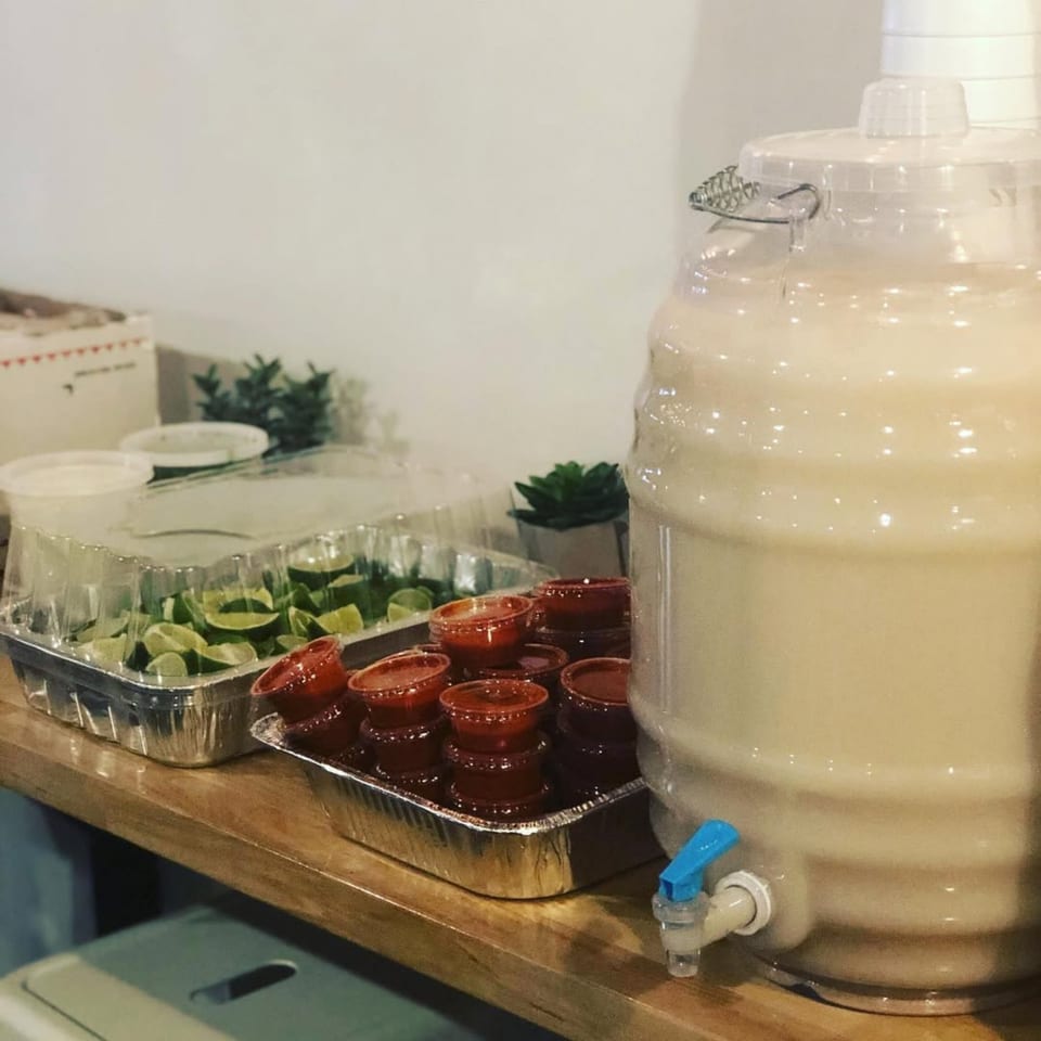A large beverage dispenser filled with horchata, alongside containers of lime wedges and red sauce.